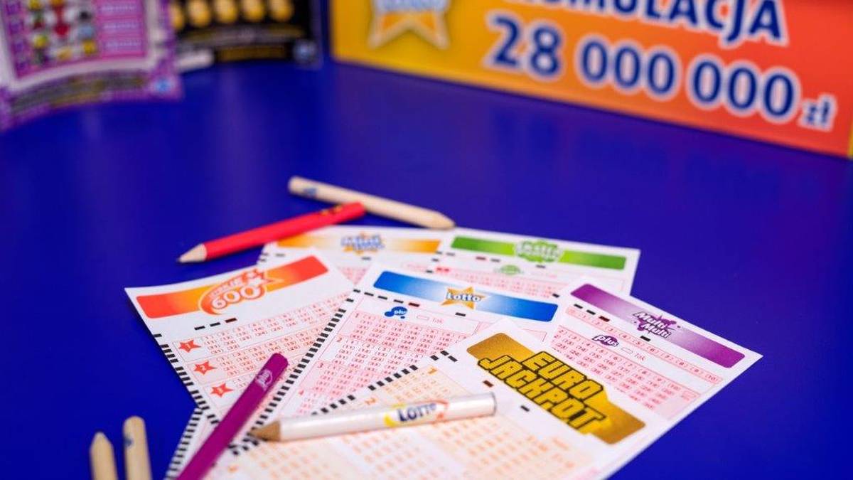 Kupony do gry Lotto i Eurojackpot leżące na niebieskim tle, obok długopis&oacute;w. W tle widoczny ż&oacute;łty baner z napisem "Kumulacja 28 000 000 zł".