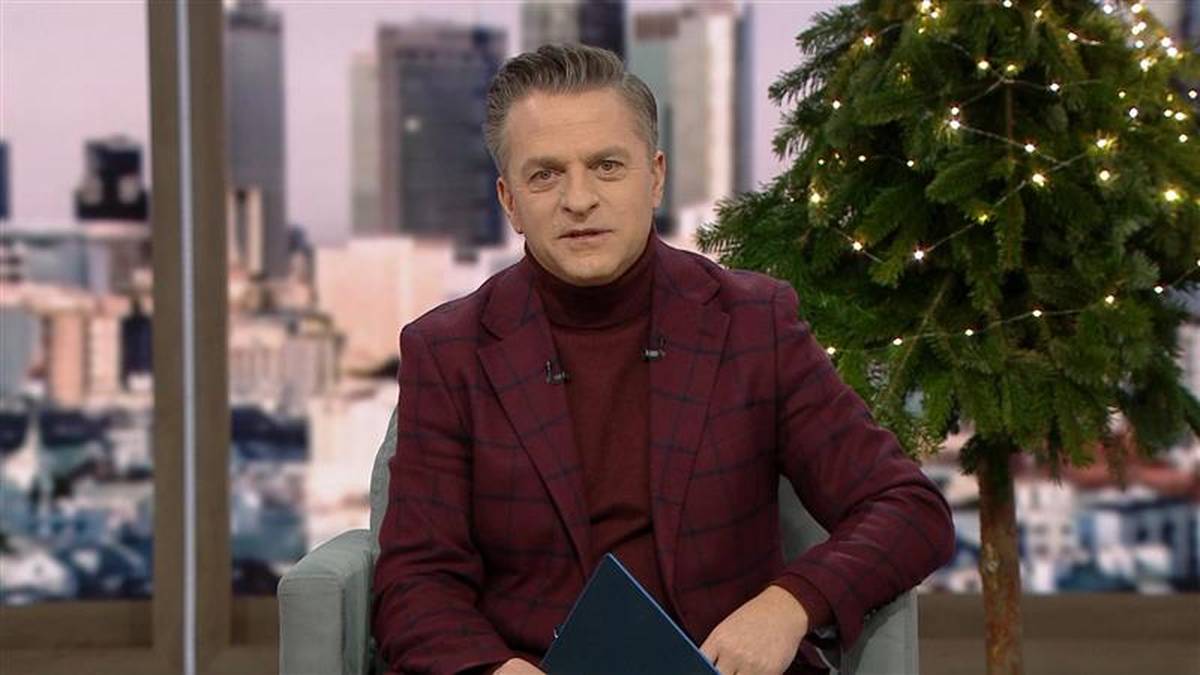 Mężczyzna w bordowym garniturze siedzi w fotelu, trzymając tablet. W tle widać panoramę miasta i choinkę ozdobioną lampkami.