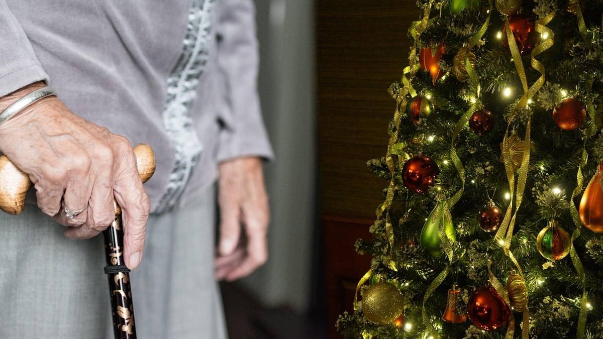 Dłoń seniora trzymająca laskę, obok choinka ozdobiona bombkami.